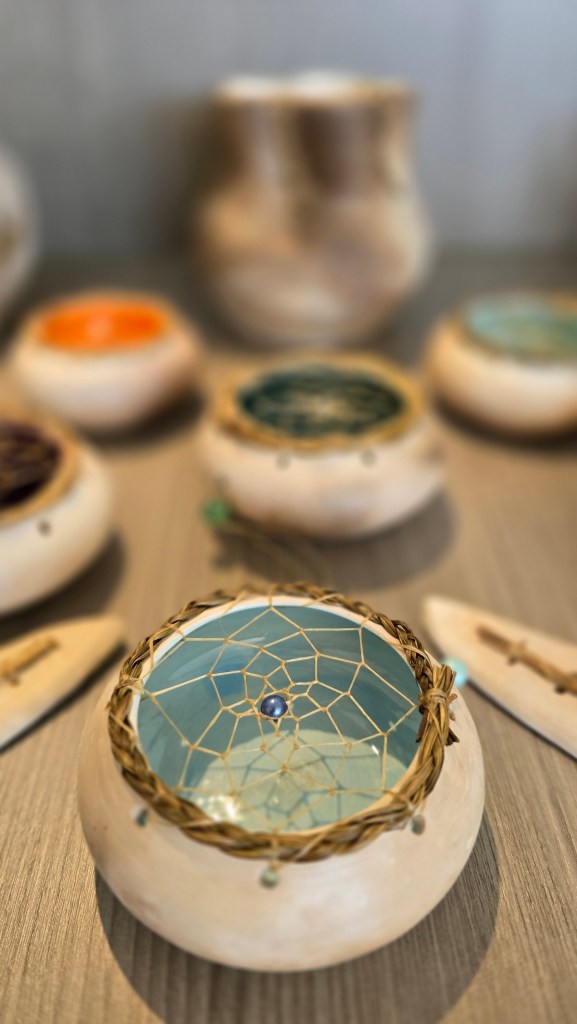 A handmade decorative bowl featuring a dreamcatcher design on top, surrounded by smaller colorful bowls and a pottery piece in the background.