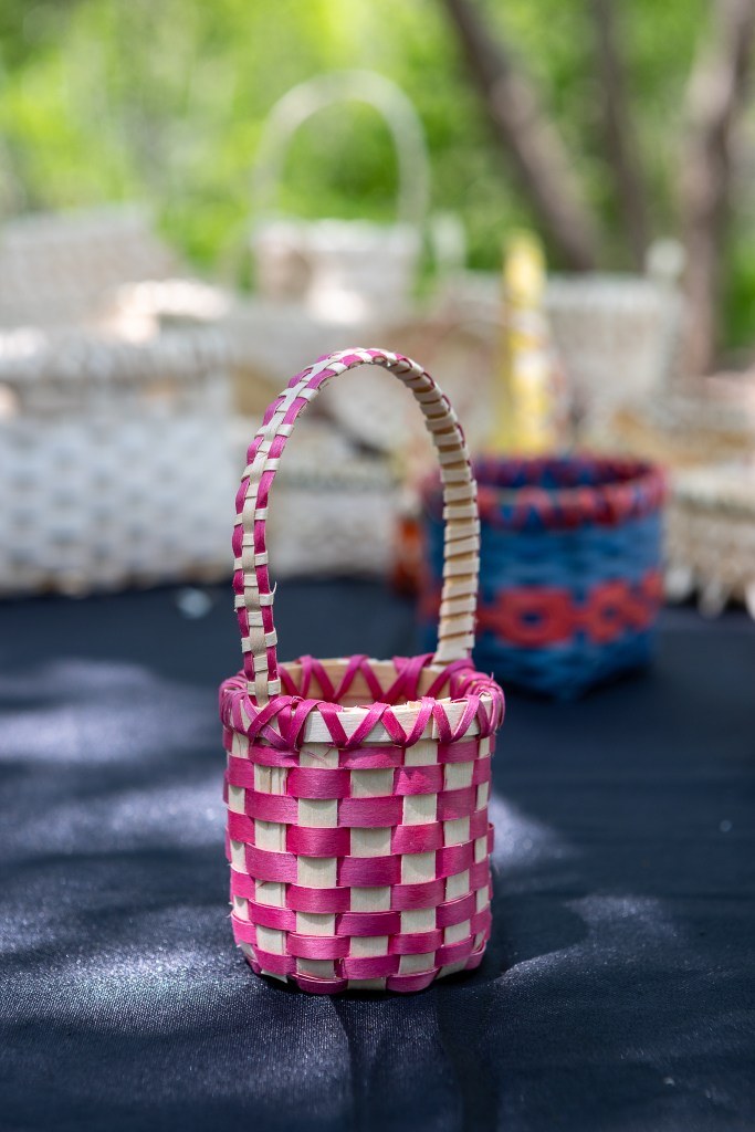 A small, handmade woven basket featuring a pink and white pattern, with a handle and surrounded by other baskets in the background.