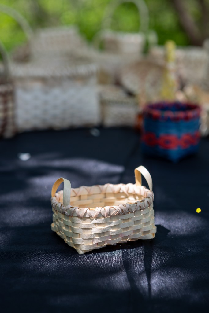 A small handmade woven basket with two handles, placed on a dark surface with other baskets blurred in the background.