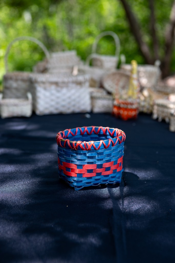 A handmade blue and red woven basket displayed on a black surface with other baskets slightly blurred in the background.