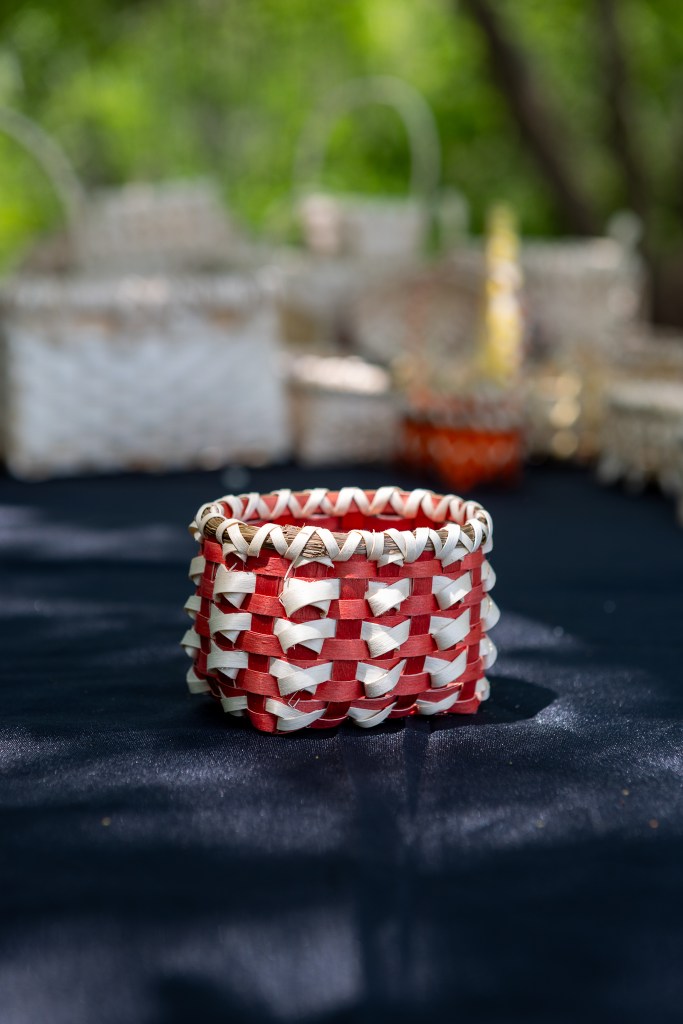 A handmade woven basket in red and white colors, resting on a dark surface with blurred basketry in the background.