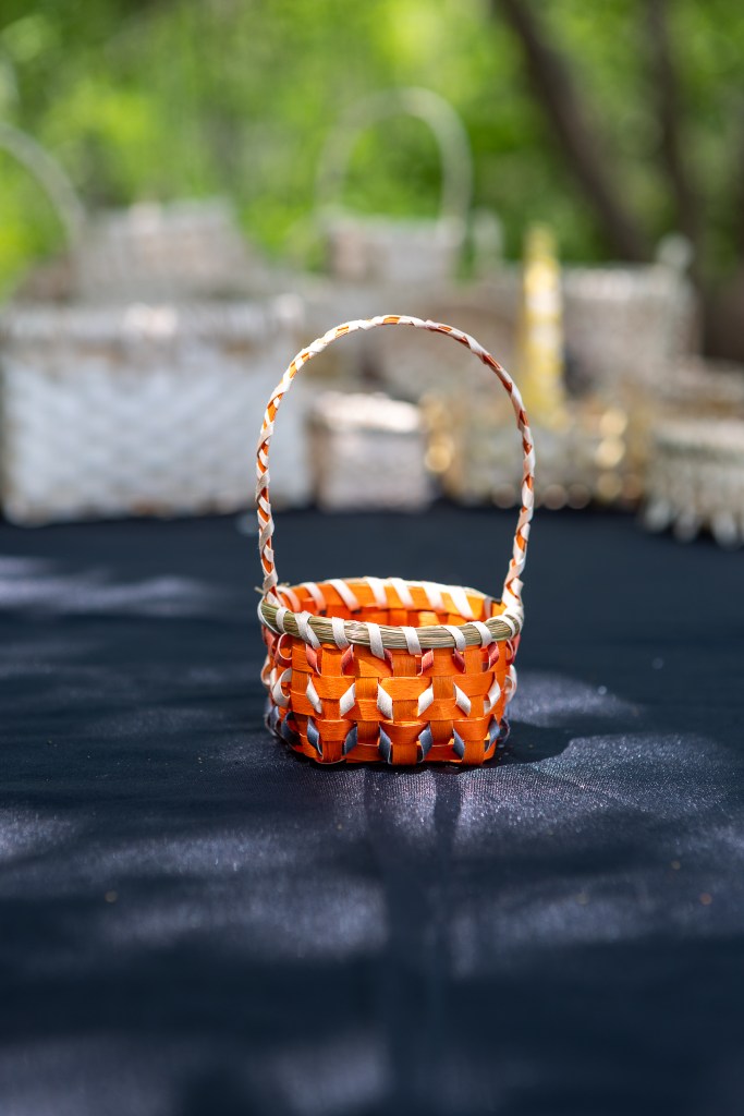 A small, brightly colored orange woven basket with a handle, sitting on a dark tablecloth, with more baskets slightly blurred in the background.
