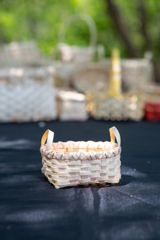 A small, handcrafted basket made of natural materials, featuring two handles, displayed on a dark tablecloth with blurred background baskets.