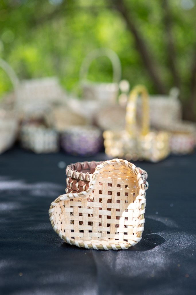 A handmade heart-shaped woven basket made by Mi'kmaq artists, displayed on a black tablecloth with other baskets in the background.