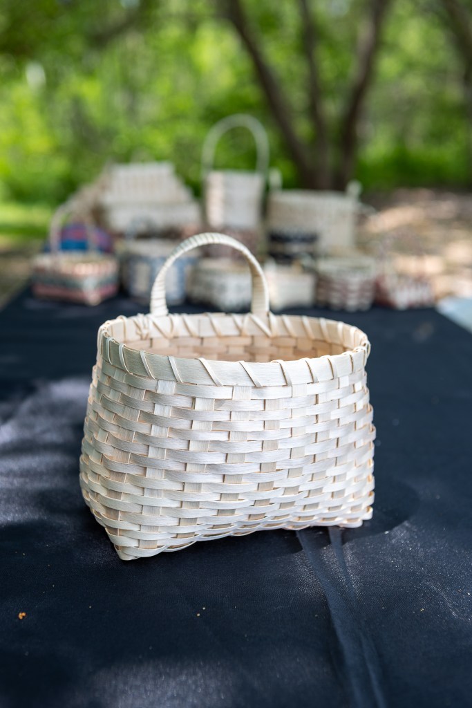 A handmade woven basket displayed on a table with more baskets in the background, surrounded by greenery.