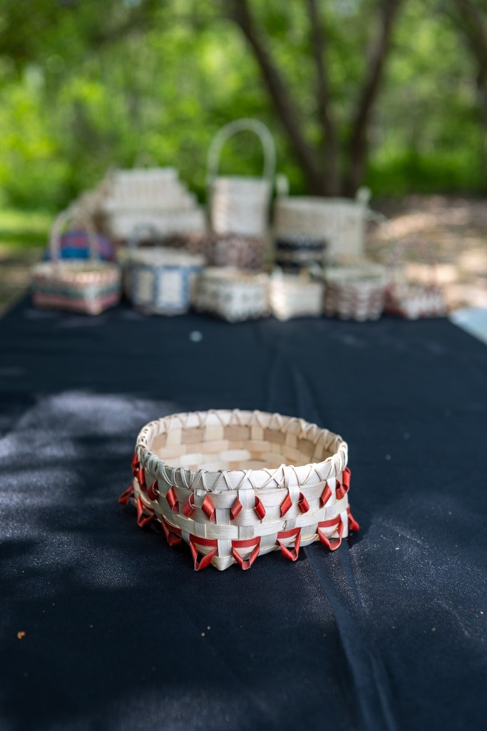 Handmade basket with red decorative accents on a black tablecloth, surrounded by other woven baskets in a natural outdoor setting.