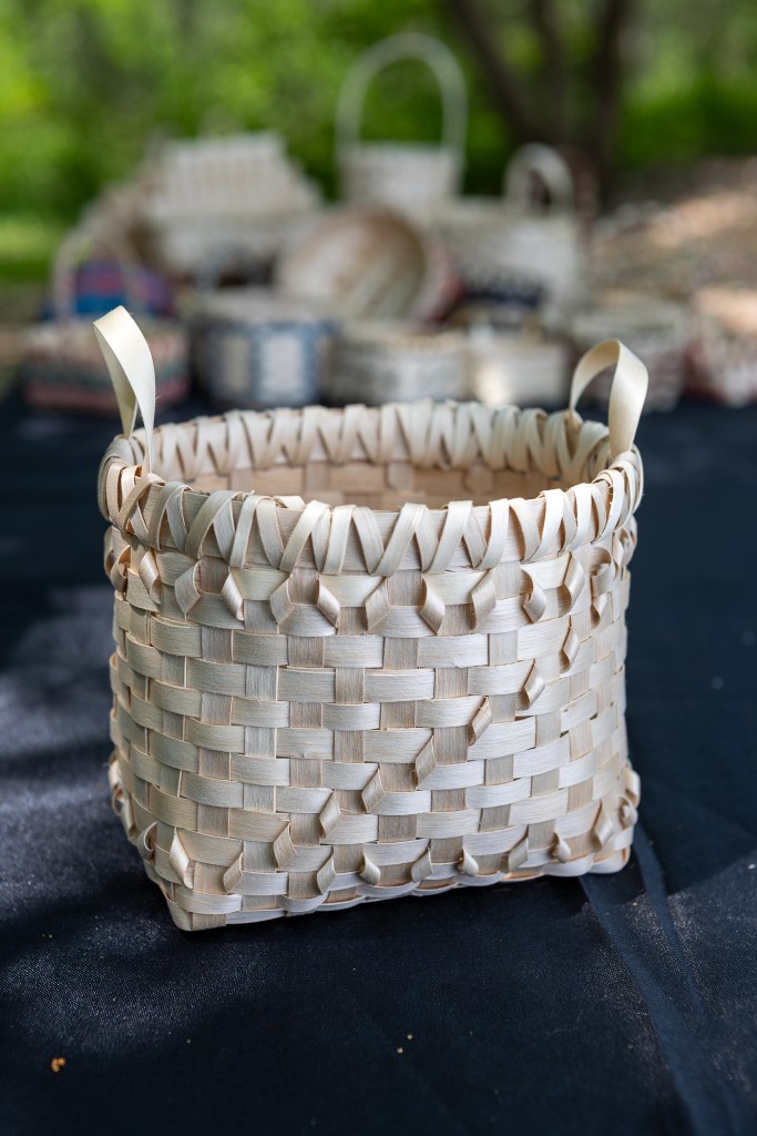A handmade woven basket with intricate patterns and handles, sitting on a dark tablecloth, surrounded by more baskets in the background.