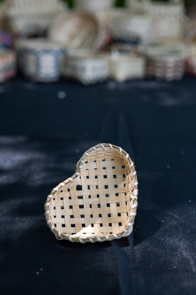 A handmade heart-shaped basket made from natural materials, placed on a dark surface with blurred woven baskets in the background.