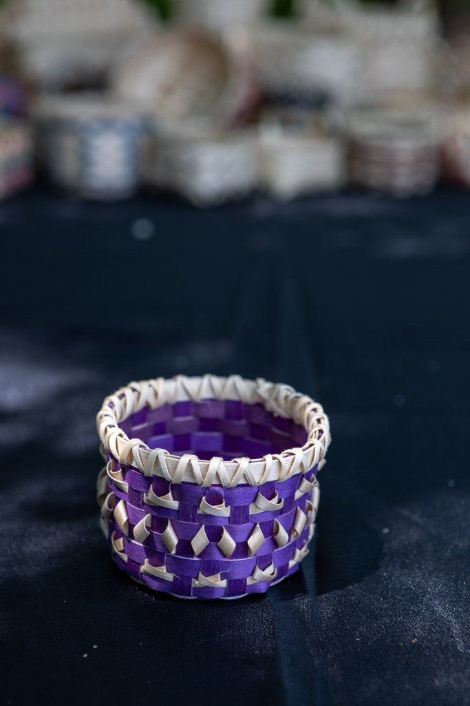 A handmade purple and cream woven basket displayed on a dark table surface, with other baskets blurred in the background.
