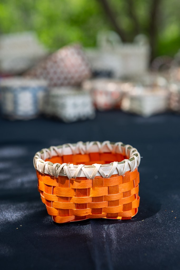 A handmade orange woven basket with a light-colored rim on a dark surface, surrounded by blurred background elements.