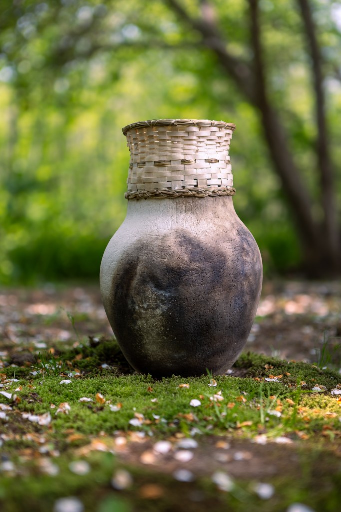 A handmade pottery vessel with a woven basket rim, resting on a bed of moss and surrounded by greenery.