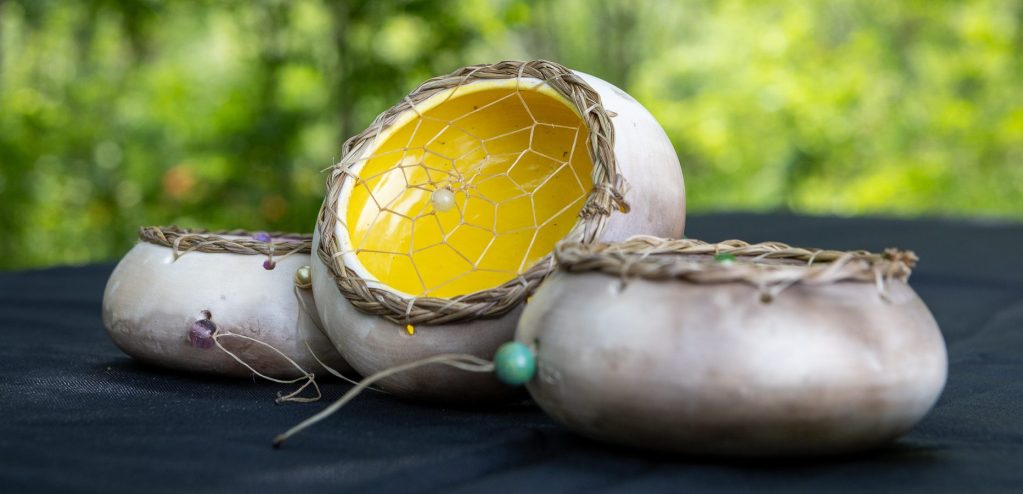 Three handmade bowls with intricate woven tops, featuring a bright yellow interior and adorned with beads, resting on a dark surface with greenery in the background.