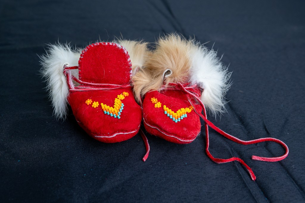 Handmade red moccasins adorned with colorful beadwork and fur trim, displayed on a dark surface.