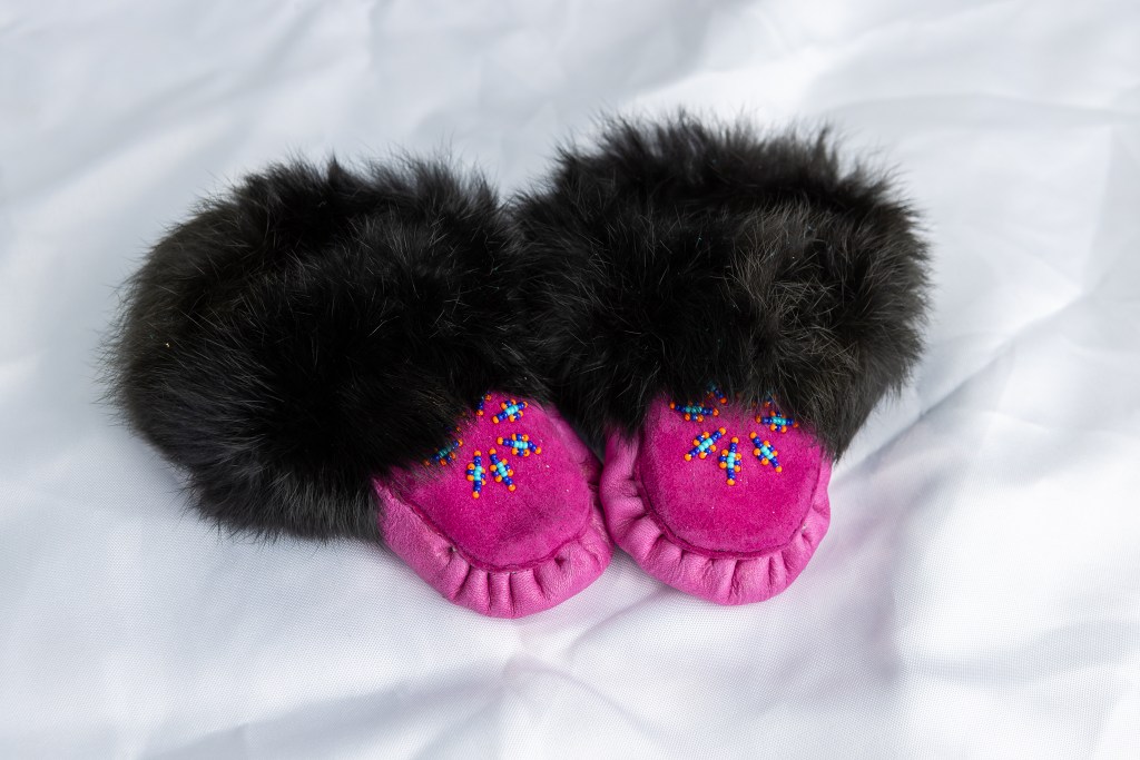 A pair of handmade Mi'kmaq moccasins with pink suede, adorned with colorful beadwork and topped with black fur trim.
